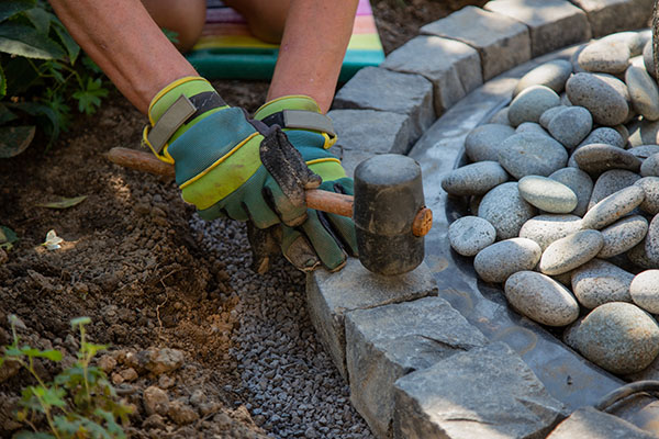 A close-up of hands in gloves using a mallet to place stones in a garden pathway, surrounded by gravel and round stones.