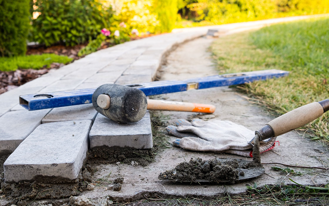 A stone pathway under construction with tools, including a mallet, level, trowel, and gloves, surrounded by greenery.