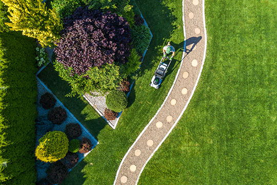 Aerial view of a person mowing a lush green lawn, surrounded by neatly trimmed bushes and a winding stone pathway.